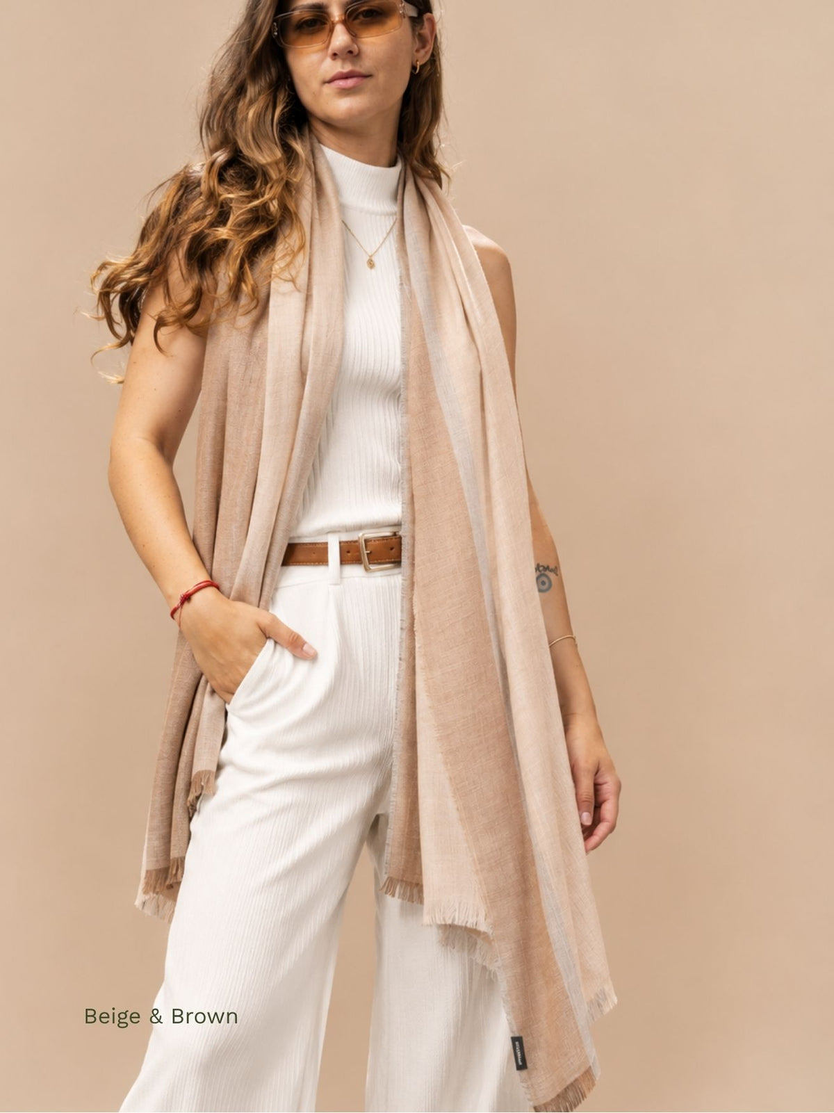 Woman wearing a beige and camel silk baby alpaca blend shawl with brown and grey stripes, draped over white trousers. Orange Inca
